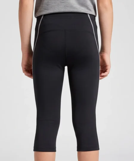 Pojkmodell visar ett par svarta knätights. Modellen visar tightsen snett bakifrån i en studio med ljusgrå bakgrund.