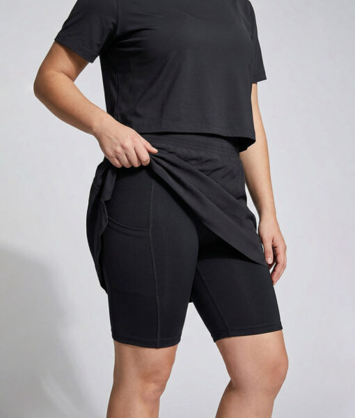 Kvinnlig modell visar en svart skort, kjol med innertights. Kvinnan lyfter upp kjolen för att visa innertightsen. Modellen står framifrån i en studio med ljusgrå bakgrund.