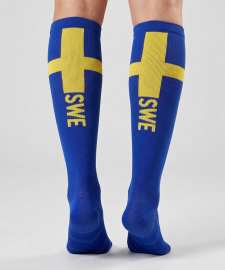 Modell visar ett par blåa och gula knähöga kompressionsstrumpor bakifrån i en studio med grå bakgrund.