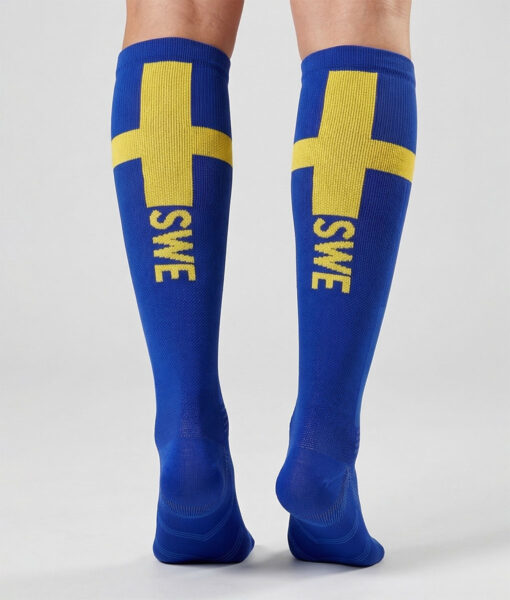 Modell visar ett par blåa och gula knähöga kompressionsstrumpor bakifrån i en studio med grå bakgrund.