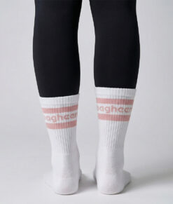 Kvinlig modell visar ett par Montana socks. Vita strumpor över svarta tights. Strumporna har rosa ränder och bagheera logo. Modellen står bakifrån i en studio med ljusgrå bakgrund.