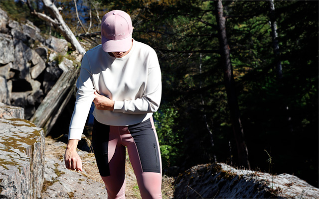 Kvinlig modell har på sig bagheera klader, benvit cloud crop top och rosa trail tights pro. Modellen vandrar i naturen. Solen skiner mellan träden.