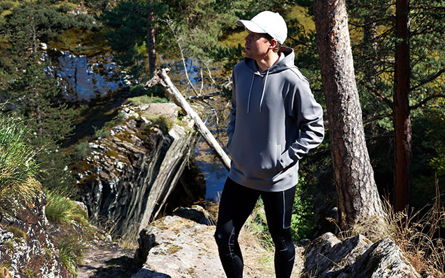 Manlig modell har på sig bagheera klader, grå cloud hoodie och svarta trail tights. Modellen står i naturen. Solen skiner mellan träden.