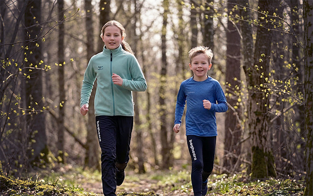 Två barn en tjej och en pojke springer i skogen. Tjejen har på sig en grån shere kahn tröja och svarta byxor. Pojken har en blå långärmad melange ls tee och svarta tights. De springer i skogen och våren är påväg.