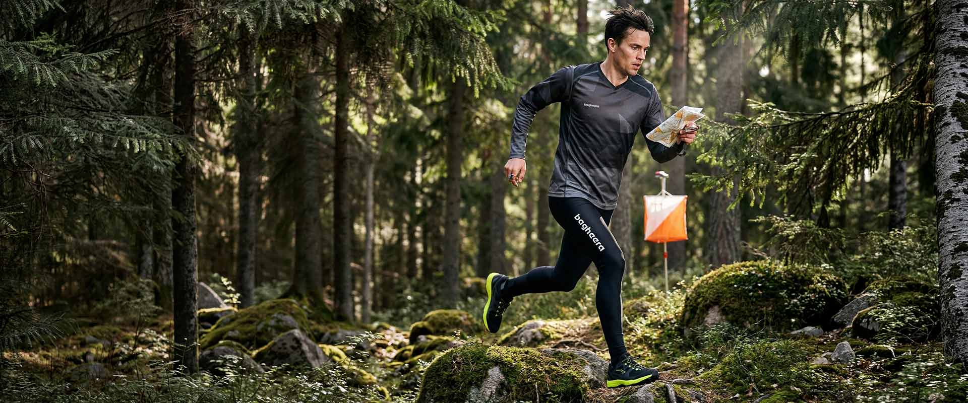 Manlig orienterare springer i tät skog med kartan i handen. Han har på sig ett par svarta tights och en svart mönstrad långärmad orienteringströja. I bakgrunden syns en orienteringskontroll.