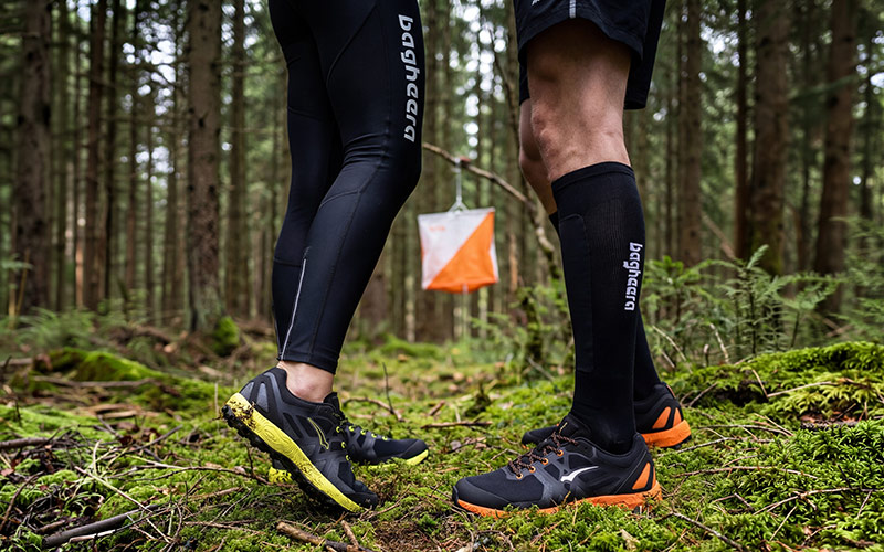 Orienteering shoes are shown in a dense forest environment. An orienteering control is visible in the background. The woman is wearing Claw X1 orienteering shoes and black tights. The guy in the picture is wearing Feral R1 orienteering shoes and black orienteering socks. The focus in the picture is on the shoes.