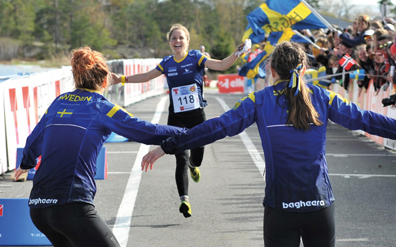 Svenska stafett orienteringslandslaget springer i mål. Tjejerna hejar på vid målgången.