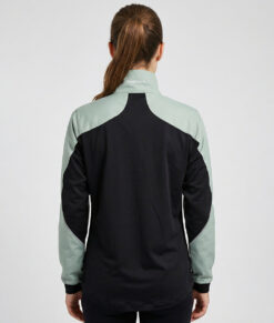 Kvinlig modell visar en ljusgrön svart vind- och vattentät träningsjacka Selva för dam i en studio med ljusgrå bakgrund. Modellen står bakifrån.