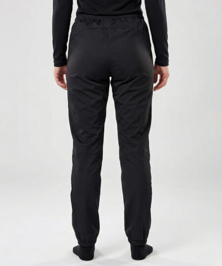 Kvinlig modell visar ett par svarta vind- och vattentäta träningsbyxor Selva för dam i en studio med ljusgrå bakgrund. Modellen står bakifrån.