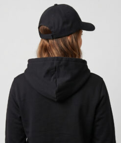 Kvinlig modell visar en svart keps. Modellen har på sig en svart o-ringen göteborg 2026 hoodie bakifrån i en studio med grå bakgrund.