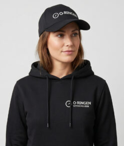 Kvinlig modell visar en svart keps med o-ringen logo. Modellen har på sig en svart o-ringen göteborg 2026 hoodie framifrån i en studio med grå bakgrund.