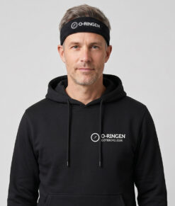 Manlig modell visar ett svart klassiskt frotépannaband med o-ringen logo. Modellen har på sig en svart o-ringen göteborg 2026 hoodie framifrån i en studio med grå bakgrund.