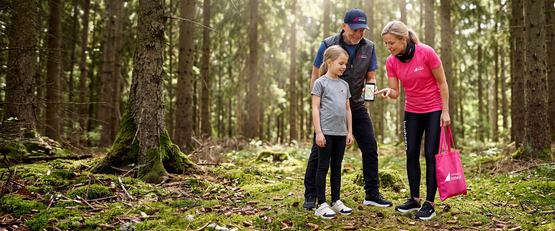 Familj hittaut orienterar i skogen iförda Bagheera kläder i hittauts kollektion. Äldre man, en kvinna och en liten tjej tittar på kartan i mobiltelefonen.