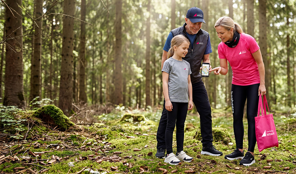 Familj hittaut orienterar i skogen iförda Bagheera kläder i hittauts kollektion. Äldre man, en kvinna och en liten tjej tittar på kartan i mobiltelefonen.