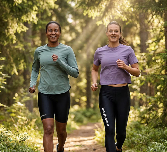 Two girls are running in the forest in bagheera clothes. One has short black tights and a green long-sleeved shirt. The other girl has long black tights and a shorter purple shirt. They are happy and the sun is shining through the trees.
