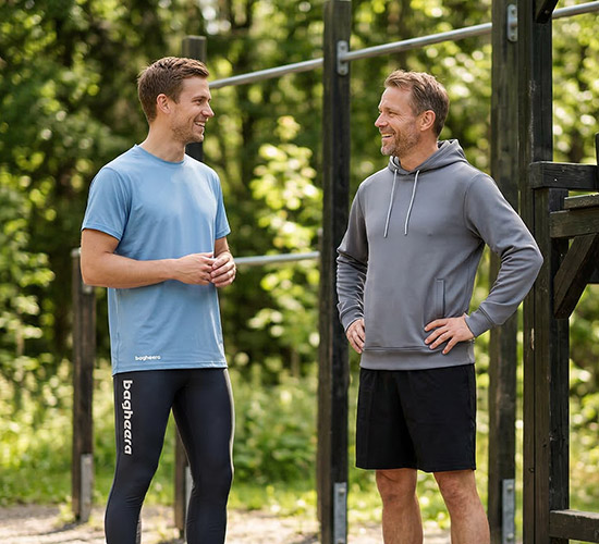 Two guys are standing and talking at an outdoor gym. One guy is wearing long black tights and a light blue t-shirt. The other guy is wearing black shorts and a gray hoodie. They are happy and the sun is shining through the trees.