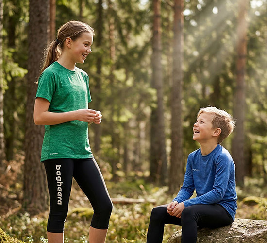 Two children, a girl and a boy, are in the forest. The sun is shining through the trees. The girl is standing up and wearing black knee-high tights and a green t-shirt. The boy is sitting on a stump, he is wearing long tights and a blue long-sleeved shirt.