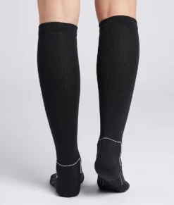 En modell visar ett par svarta knähöga compressionsstrumpor i varm merinoull. Modellen står bakifrån i en studio med ljusgrå bakgrund.