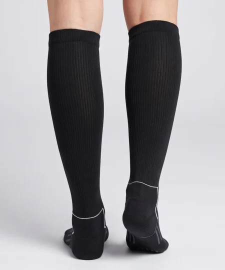 En modell visar ett par svarta knähöga compressionsstrumpor i varm merinoull. Modellen står bakifrån i en studio med ljusgrå bakgrund.