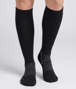En modell visar ett par svarta knähöga compressionsstrumpor i varm merinoull. Modellen står framifrån i en studio med ljusgrå bakgrund.