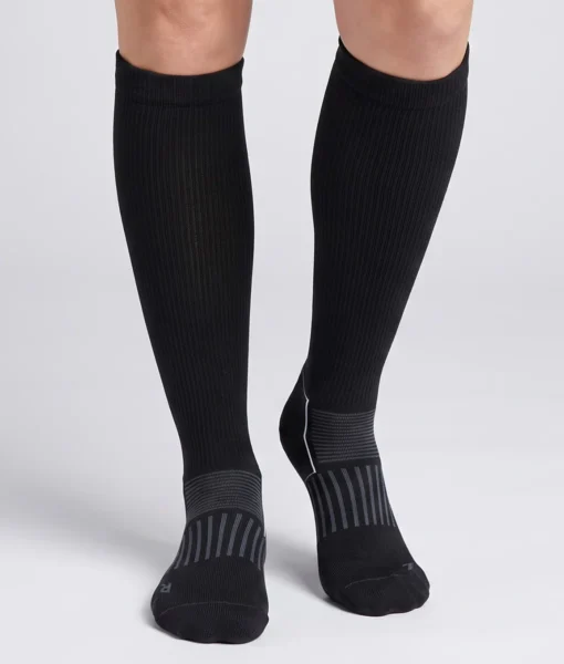 En modell visar ett par svarta knähöga compressionsstrumpor i varm merinoull. Modellen står framifrån i en studio med ljusgrå bakgrund.