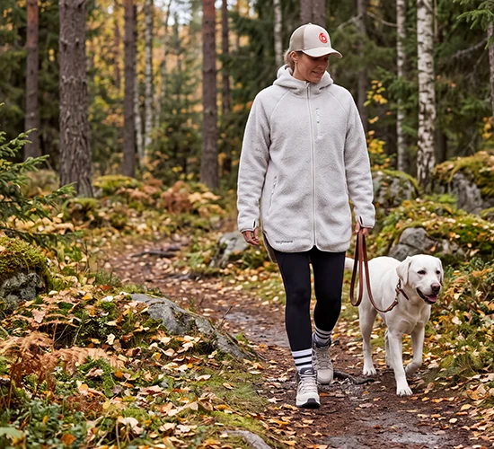 En kvinna rastar sin vita labrador i en höstig skog. Kvinnan har på sig en beige trucker keps, en vit commfy hood jacket, svarta tights och ett par hiker skor från bagheera.