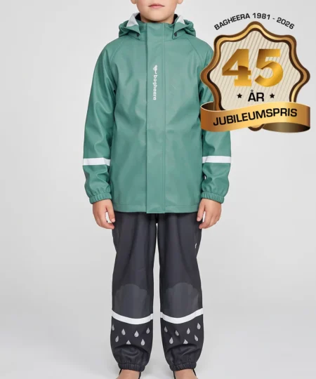 Pojkmodell visar en dimblå regnjacka och svarta regnbyxor i ett paketpris. Modellen står framifrån i en studio med ljusgrå bakgrund.